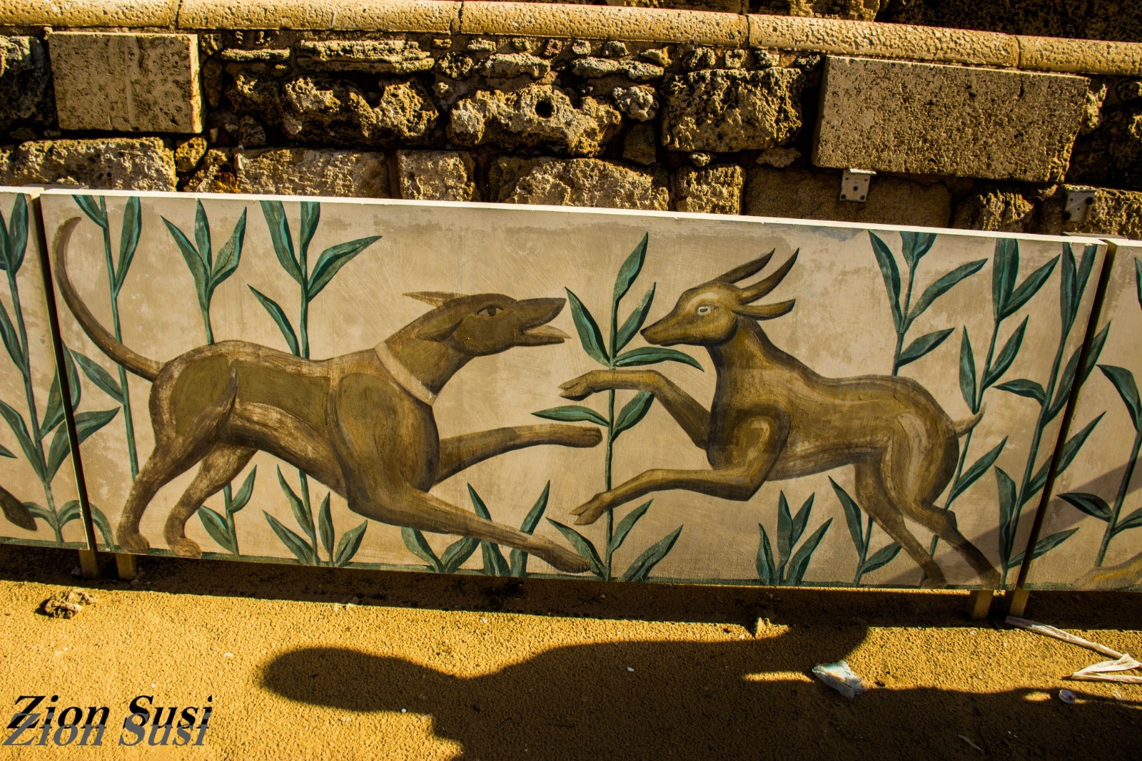 Stylized mural of a dog and gazelle among green plants on a weathered stone wall at Caesarea