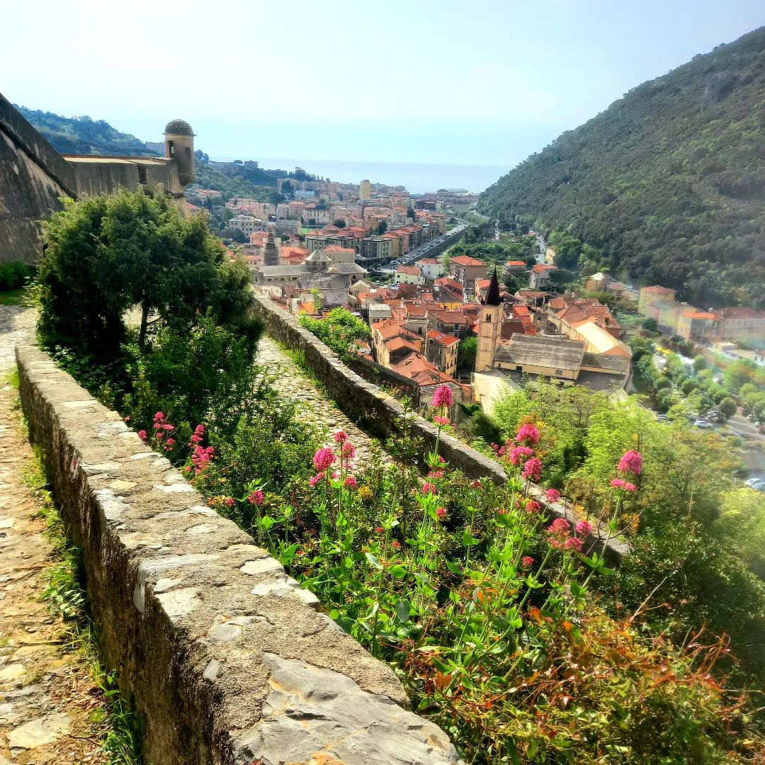 View of Finalborgo from the Castel San Giovanni path with flowering stone wall and Ligurian hills